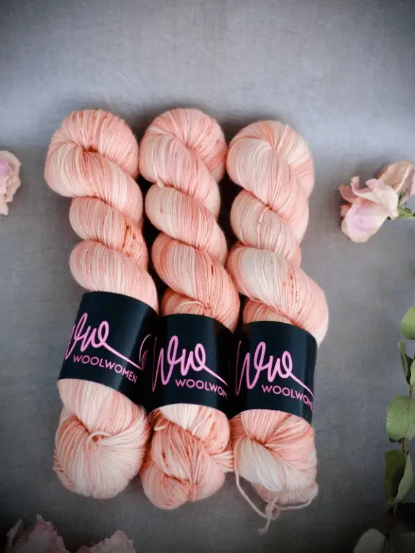 Three skeins of pale peach WoolWomen Merino Sock yarn are laid side by side on a gray surface, surrounded by soft pink flowers and green leaves.