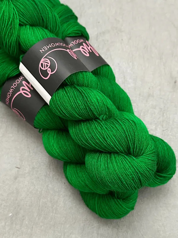 Two skeins of vibrant green WoolWomen Merino Sock yarn are stacked on a gray surface, each käsinvärjätty lanka wrapped with a black label featuring pink and white text and circular logos.