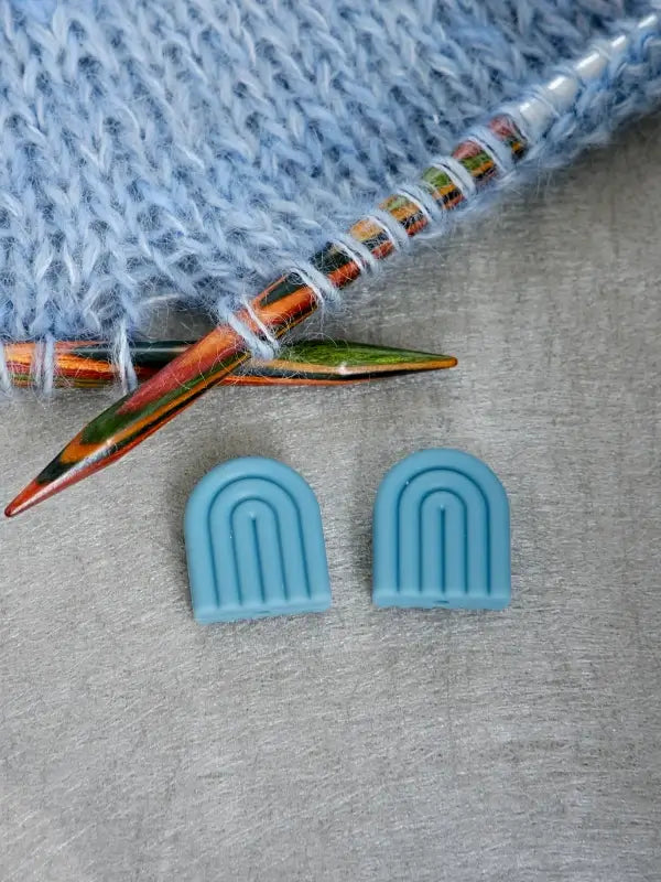 Two colorful knitting needles inserted in blue yarn are placed beside two small, blue, arch-shaped Woolwomen Puikkostopparit needle stoppers on a gray surface.
