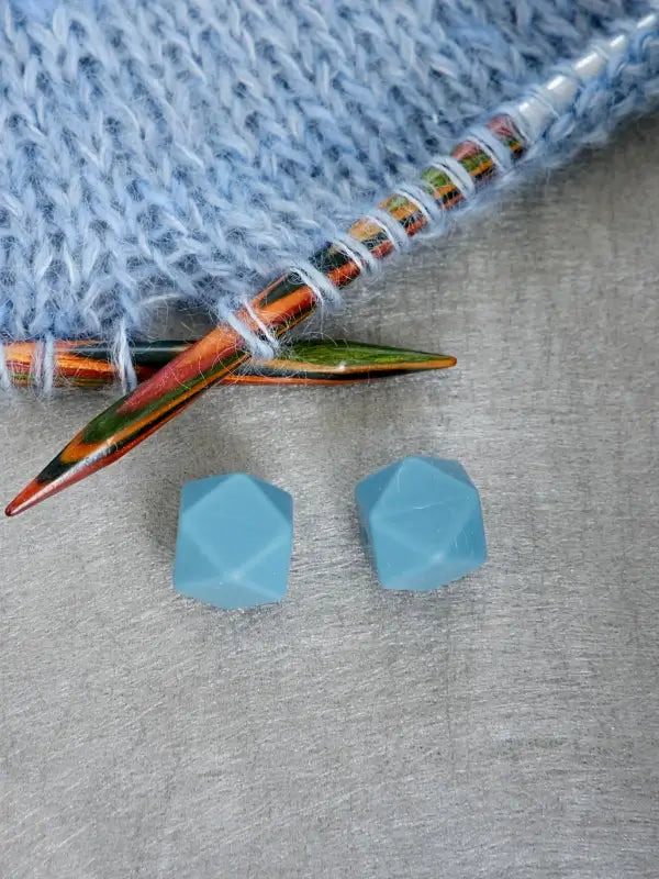 Two faceted, light blue, hexagonal Woolwomen Puikkostopparit rest on a gray surface beside colorful knitting needles and light blue knitted fabric.