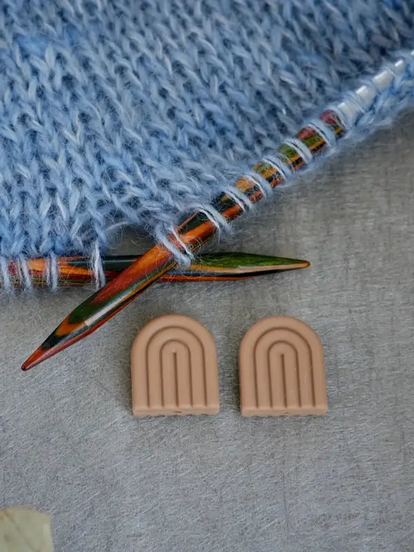 Colorful knitting needles hold blue yarn. Nearby, two small, beige Woolwomen Puikkostopparit in the shape of rainbows rest on a light gray surface.