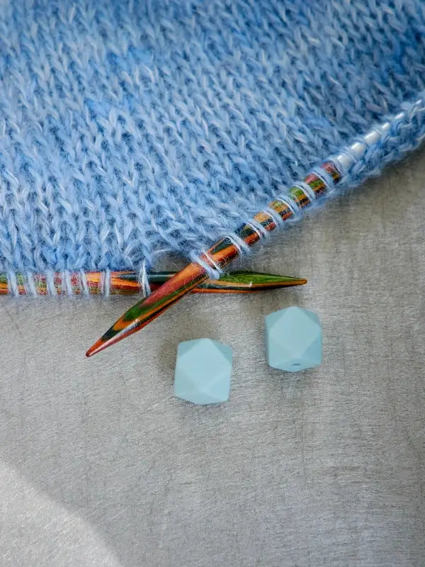 Woolwomen Puikkostopparit shown with blue yarn forming fabric on knitting needles, placed next to two faceted light blue beads on a gray surface.