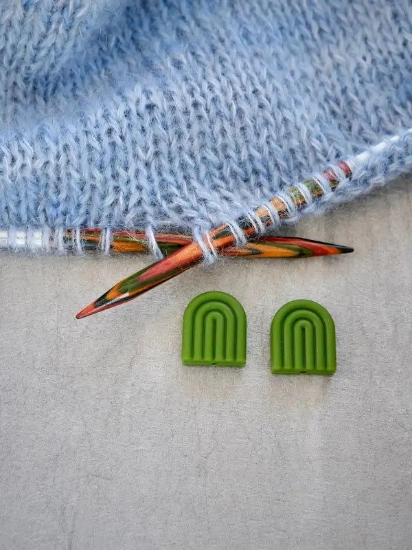 A close-up of multicolored knitting needles working on light blue yarn, with two small, green Woolwomen Puikkostopparit placed on a light surface below.