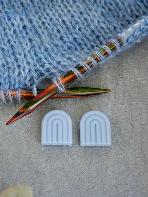 Two colorful knitting needles rest on a soft blue knitted fabric. Below them are two small, white, rainbow-shaped Woolwomen Puikkostopparit on a light gray surface.