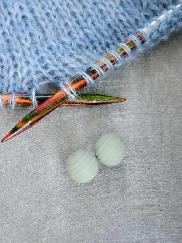 Two green, round Woolwomen Puikkostopparit rest on a gray surface next to colorful wooden knitting needles and a piece of blue knitted fabric.