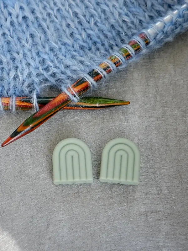 Two wooden knitting needles with light blue yarn and two light green Woolwomen Puikkostopparit arch-shaped needle point protectors on a gray surface.
