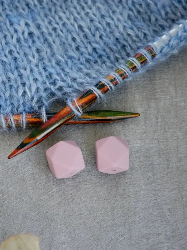 A pair of Woolwomen Puikkostopparit—pink, faceted knitting needle stoppers—rest on a gray surface beside colorful knitting needles inserted into light blue yarn.