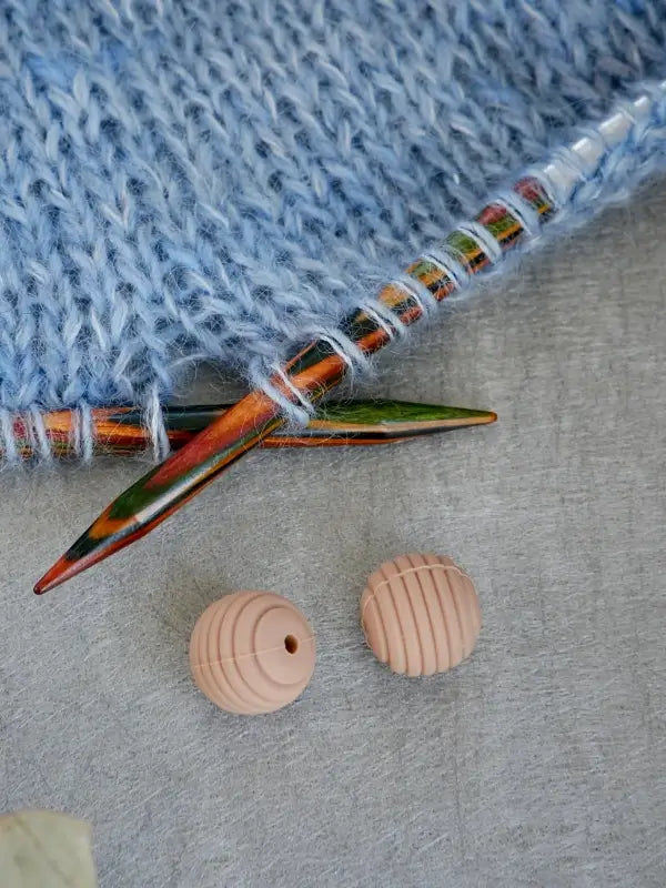 Two multicolored knitting needles hold blue yarn on a light gray surface, with two round, ribbed Woolwomen Puikkostopparit in light beige nearby.