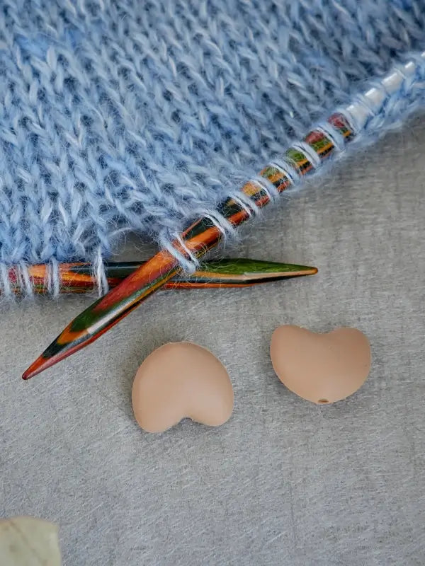 Woolwomen Puikkostopparit knitting needles with light blue yarn and two small, heart-shaped beige buttons resting on a gray surface.
