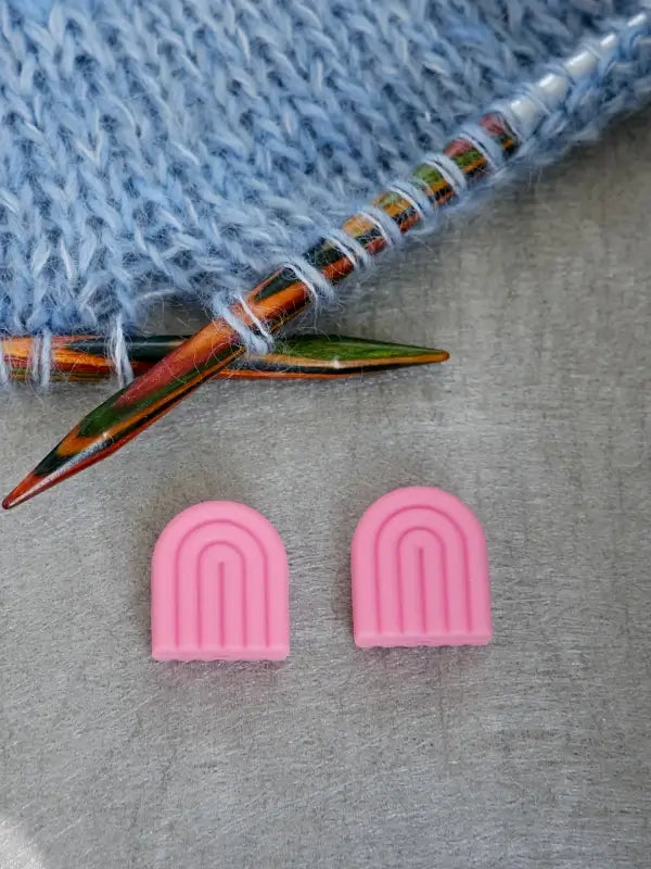 Two colorful knitting needles hold light blue yarn, and below them on a gray surface are two small, pink Woolwomen Puikkostopparit—arch-shaped silicone needle stoppers with a rainbow-like design.