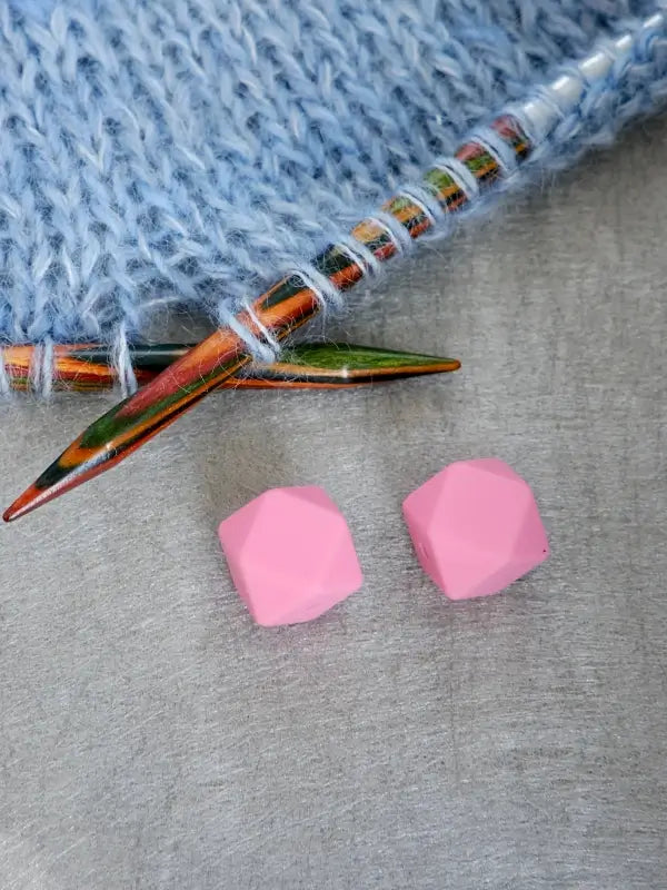 Two pink, faceted hexagonal Woolwomen Puikkostopparit rest on a gray surface near two colorful wooden knitting needles and light blue knitted fabric.