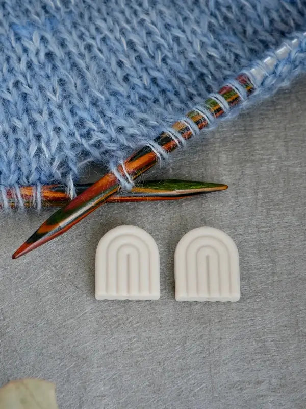 Two Woolwomen Puikkostopparit white rainbow-shaped stitch stoppers rest on a gray surface beside colorful wooden knitting needles and blue knitted fabric.