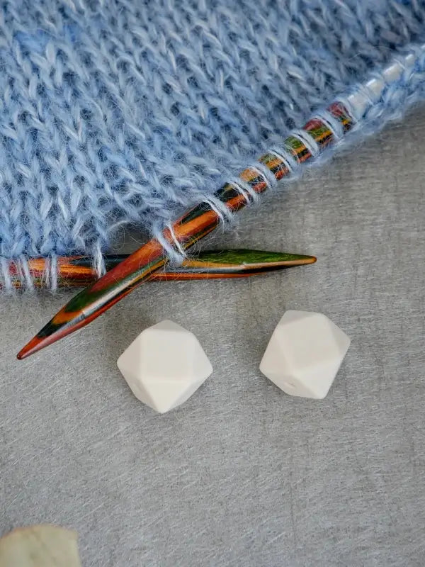 A close-up of the Woolwomen Puikkostopparit wooden knitting needles with blue yarn, displayed next to two white geometric beads on a gray surface.