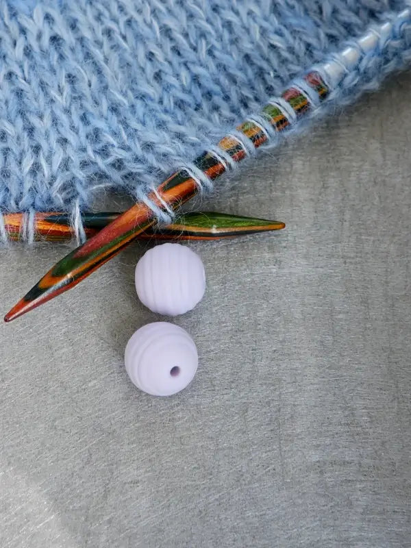 Two colorful wooden knitting needles are inserted in a piece of light blue knitting. Below them, two round, textured, pale purple Woolwomen Puikkostopparit rest on a gray surface.