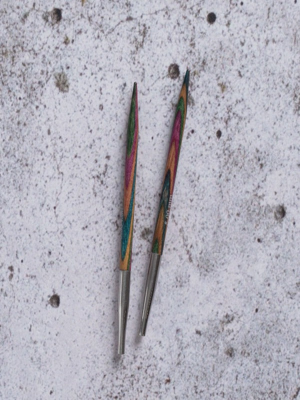 Two colorful wooden knitting needle tips with metal bases are placed on a speckled, light gray surface.