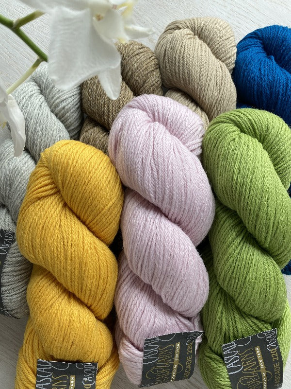 Several skeins of yarn in gray, yellow, pink, green, beige, and blue are arranged on a light surface. A white flower is partially visible in the top left corner of the image.