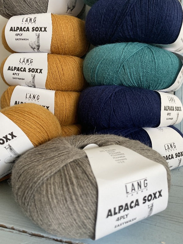 Several skeins of Lang Alpaca Soxx yarn in mustard yellow, teal, navy blue, and gray are stacked together on a wooden surface. Each skein has a white label.