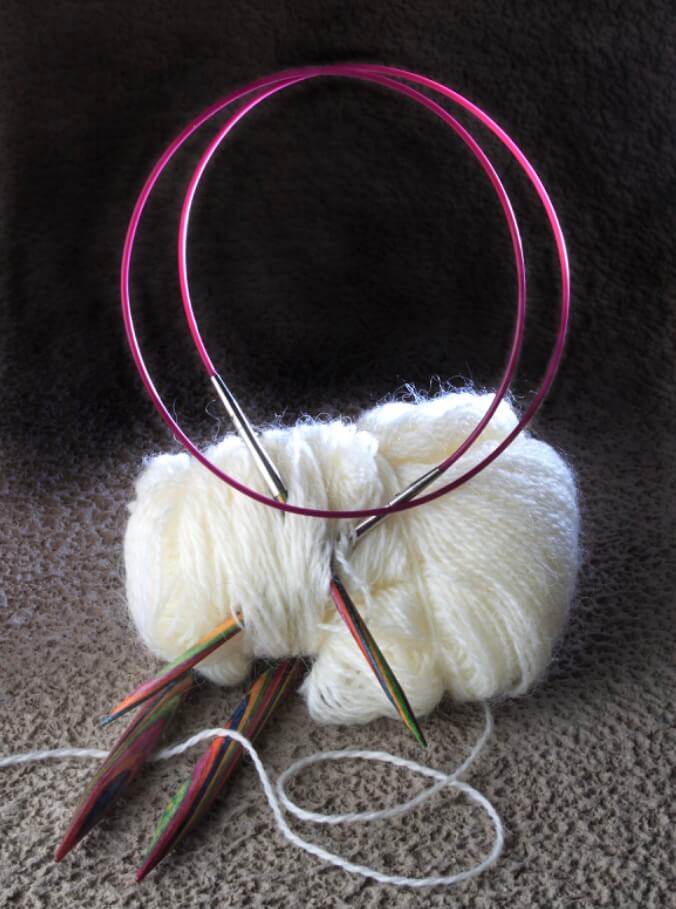 A skein of white yarn with two colorful circular knitting needles inserted into it, the pink cables looped above on a textured brown surface.