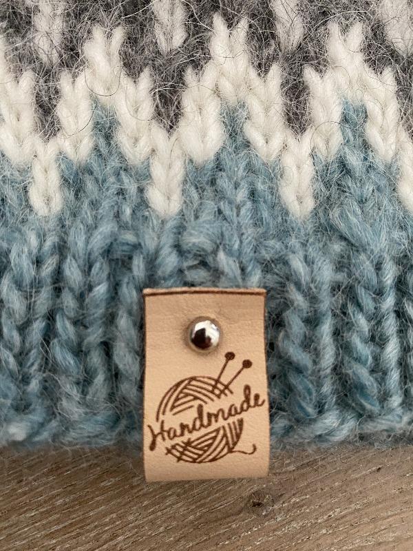 A close-up of knitted fabric in grey, white, and blue tones with a leather tag that reads Handmade and features a yarn ball with knitting needles.