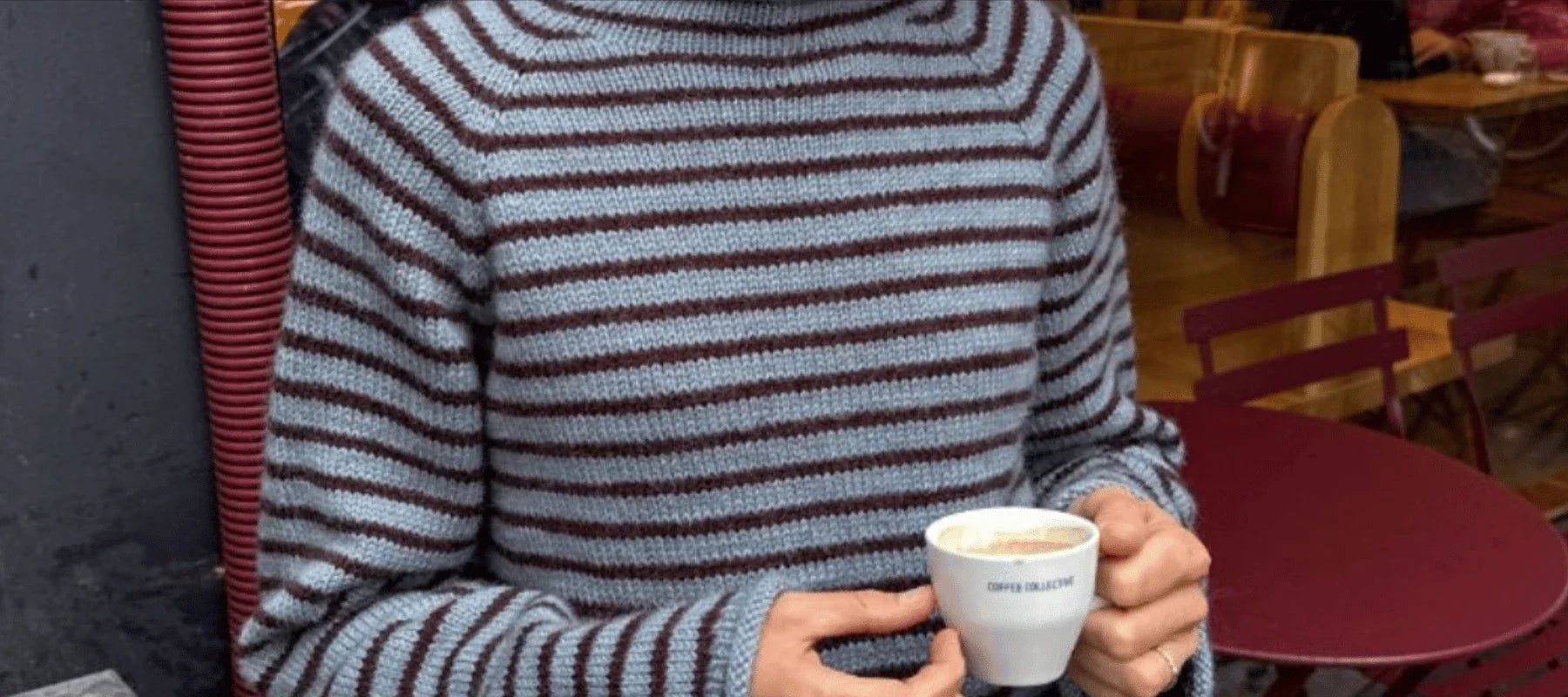 A person in a blue and brown striped turtleneck sweater holds a small cup of coffee while sitting at a round red table in a cozy café.