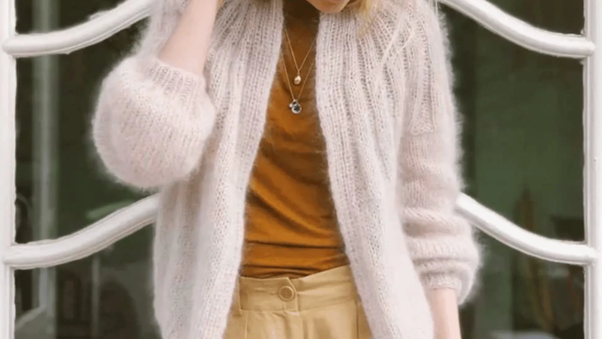 A person wearing a fuzzy cream cardigan, a rust-colored top, and tan high-waisted pants stands in front of a white, curved metal structure, with one hand touching their hair.