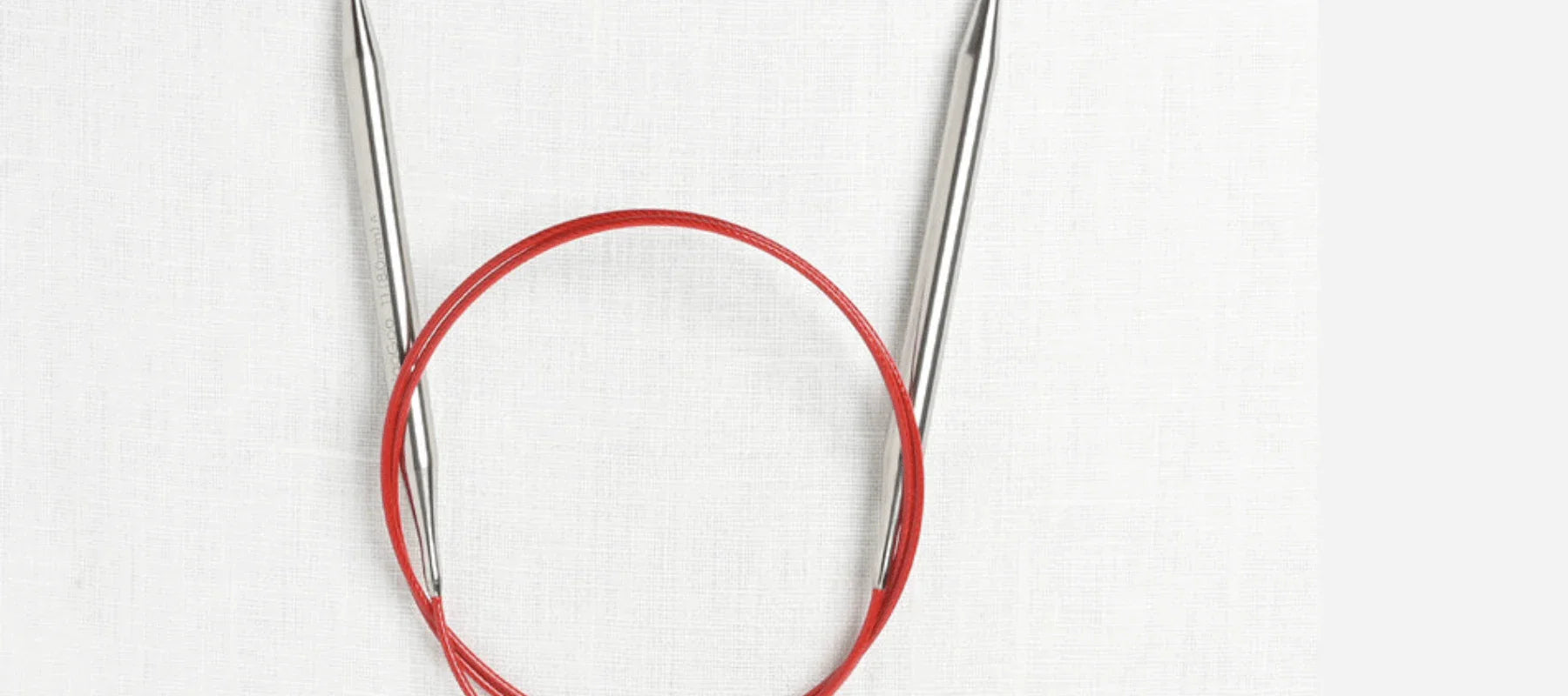A pair of metal circular knitting needles connected by a flexible red cable, laid out on a white textured background.