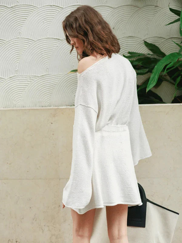 A woman with wavy brown hair wears an off-shoulder white knit dress with long sleeves, standing indoors near a textured wall and green plant, facing away from the camera.