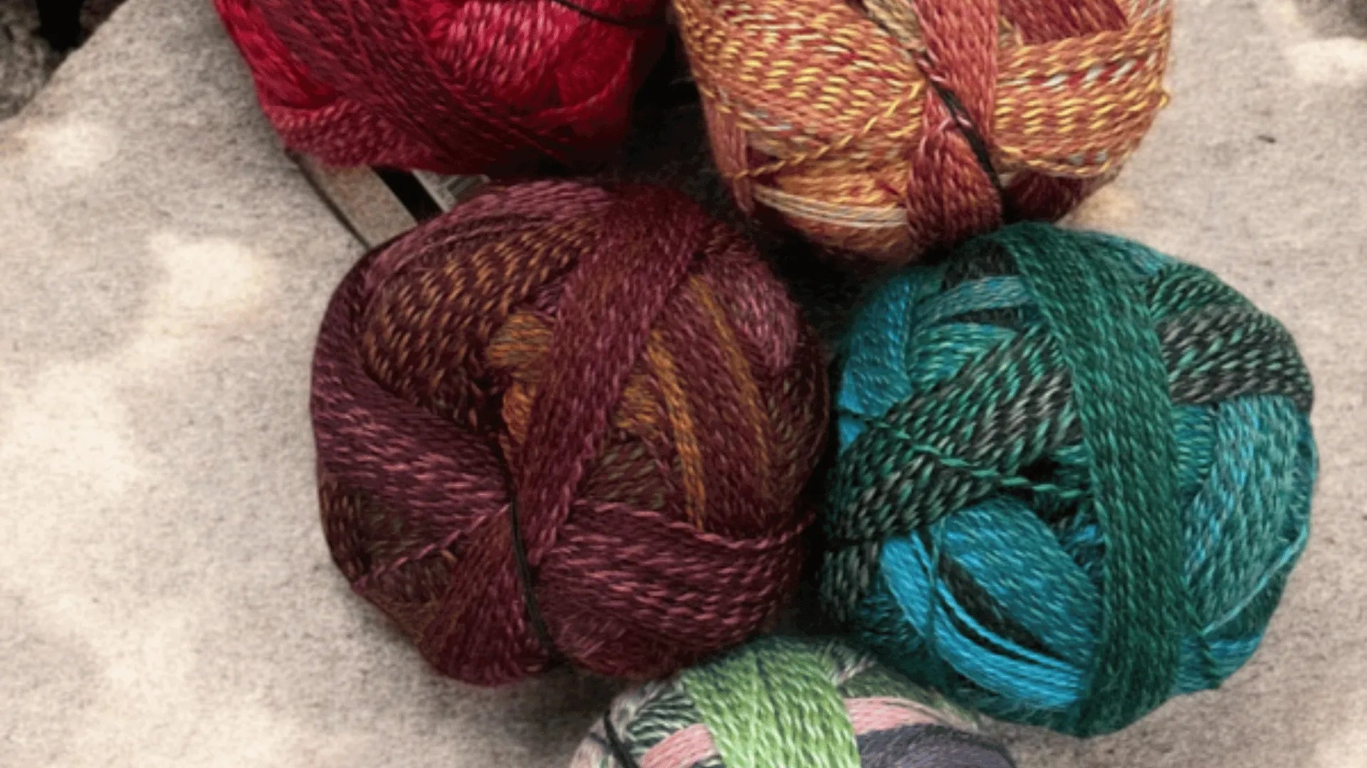 Five balls of multicolored yarn—red, gold, burgundy, green, and blue—are arranged on a light, textured surface.