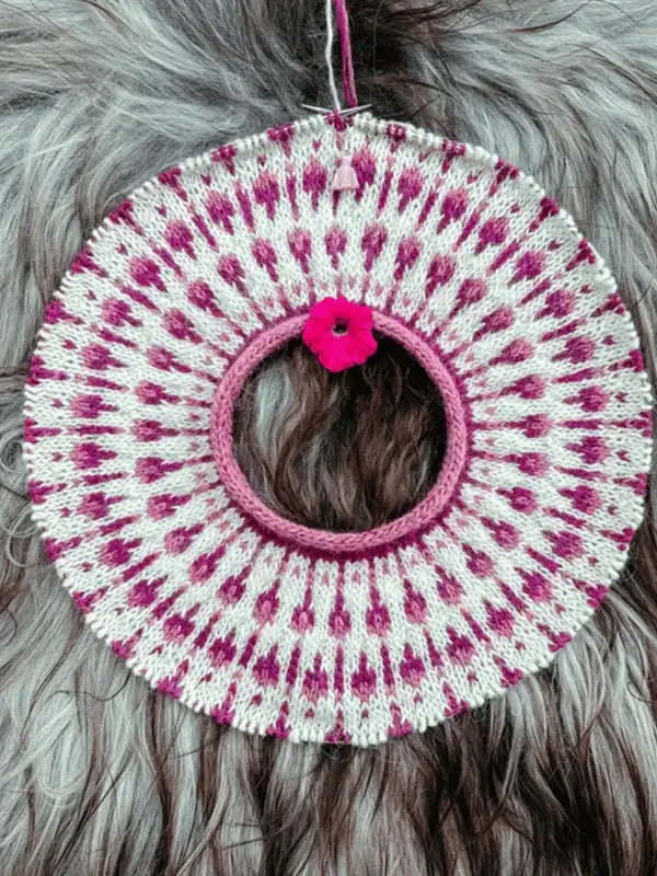 A circular, knitted piece with pink and white patterns, resembling a decorative collar. It has a pink flower attached to one side and is placed on a furry, gray and brown background.