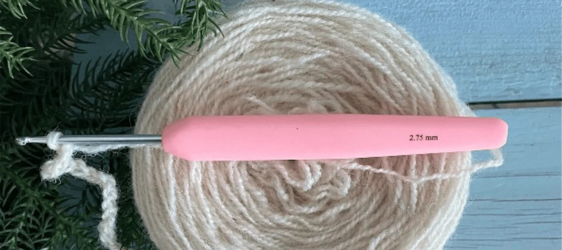 A pink-handled 2.75 mm crochet hook rests on a cream-colored ball of yarn, with a green plant and light blue wooden surface in the background.