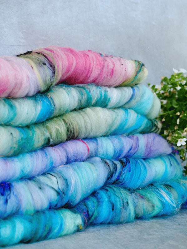 Four stacked bundles of hand-dyed wool in pastel shades of blue, pink, and green, with a small plant with white flowers in the background.