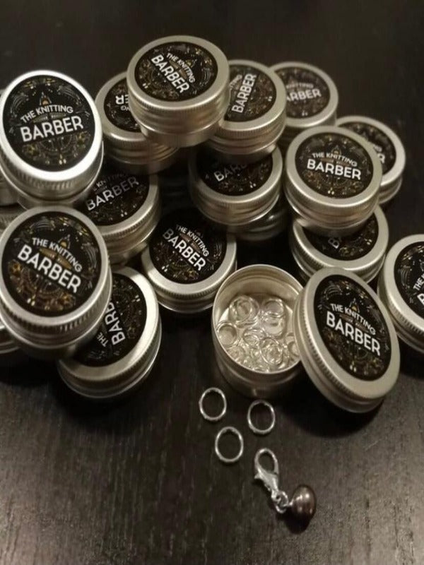 Several small metal tins labeled The Knitting Barber are stacked on a dark surface. Some tins are open, revealing knitting stitch markers and a clasp inside.