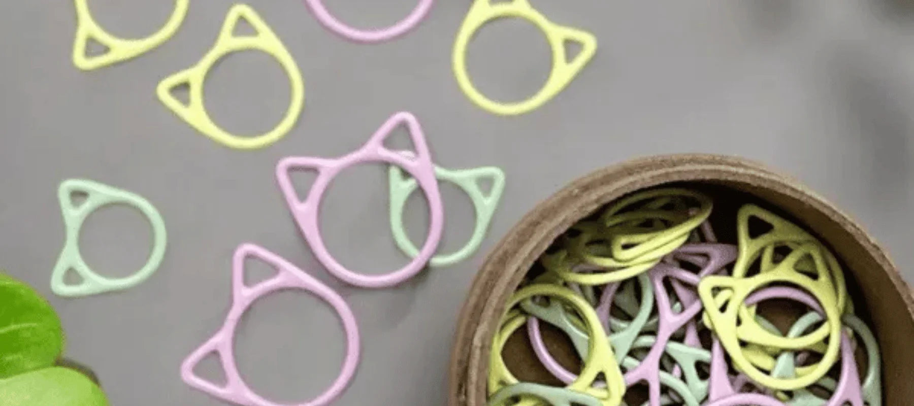 Pastel-colored cat-shaped paper clips in yellow, green, and purple are scattered on a gray surface, with more clips inside a round wooden container.