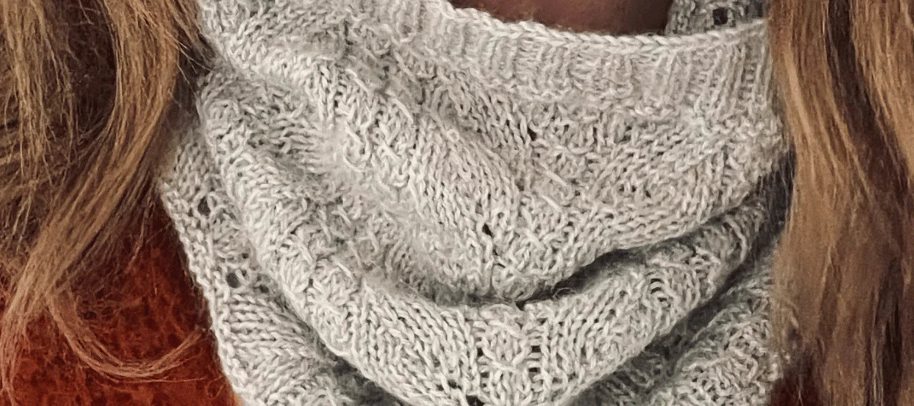 Close-up of a person wearing a soft, light gray knitted scarf with a textured pattern. The person has light brown hair and is also wearing a rust-colored sweater.