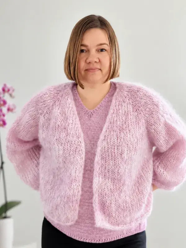 A woman with straight, light brown hair wears a soft, fluffy pink cardigan made from WoolWomen Fluff Mohair, knitted using the Fluff neuletakki ohje pattern, over a pink top. She stands indoors near a potted orchid against a light background.