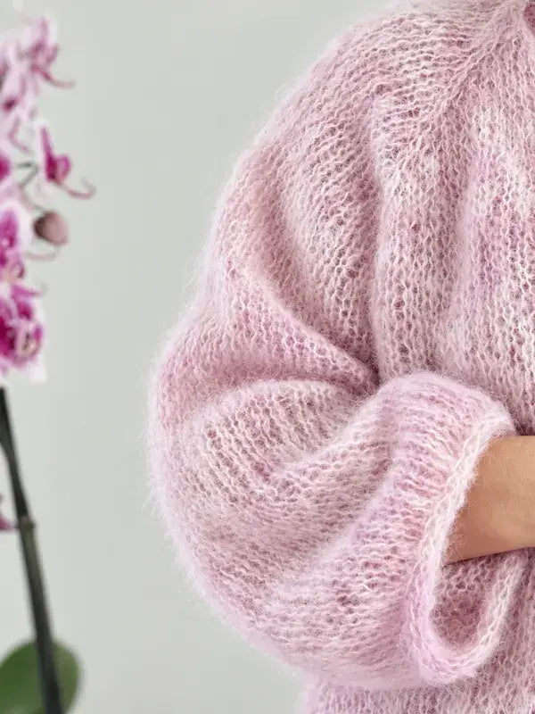 A person wearing a soft, fluffy pink Fluff neuletakki ohje with rolled-up sleeves stands beside a blooming orchid plant.