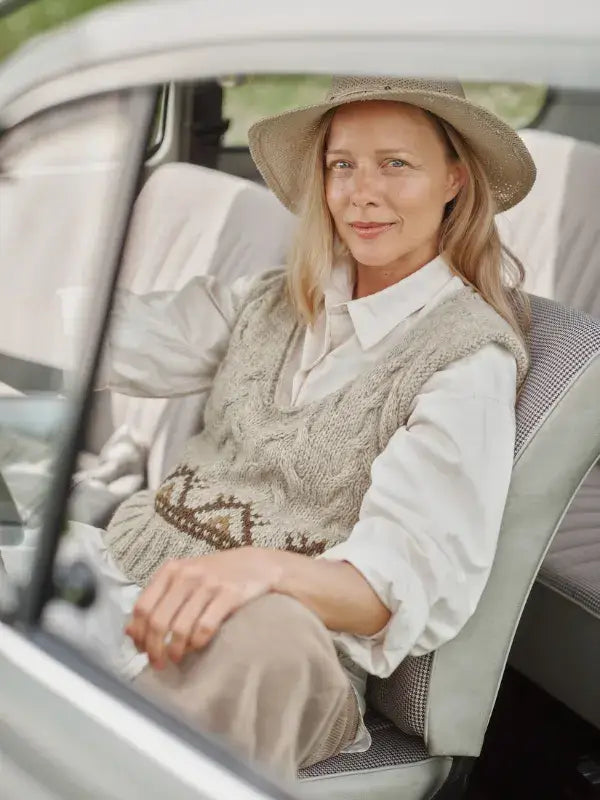 A woman in a straw hat and a patterned Jenna Kostet - Neulottu Kalevala III, Ennakkotilaus knit vest sits in the driver’s seat of a car.