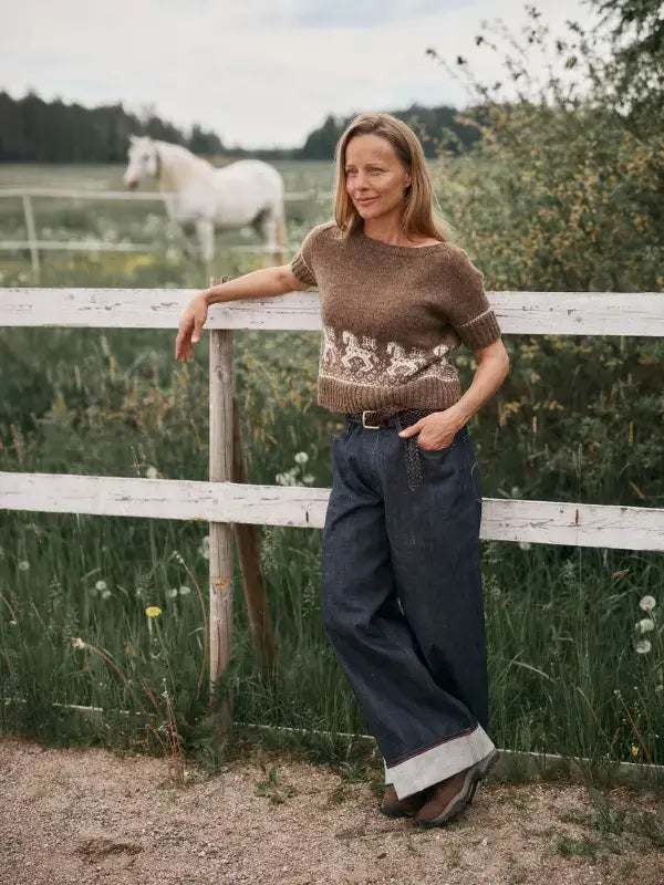 A person wearing a brown Neulottu Kalevala III sweater with a white horse pattern leans against a white wooden fence in a field with a horse in the background.