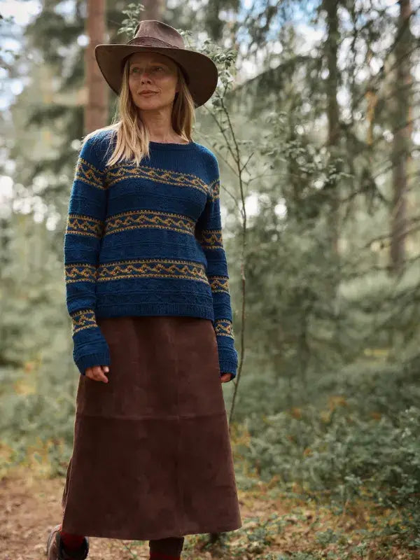 A woman in a brown hat and a Jenna Kostet - Neulottu Kalevala III, Ennakkotilaus knit sweater over a brown skirt stands in a forest.