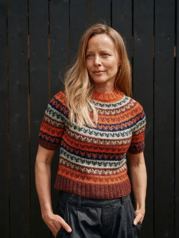 A woman wearing a Jenna Kostet - Neulottu Kalevala III, Ennakkotilaus short-sleeved sweater featuring a multi-colored geometric Fair Isle kirjoneule pattern.