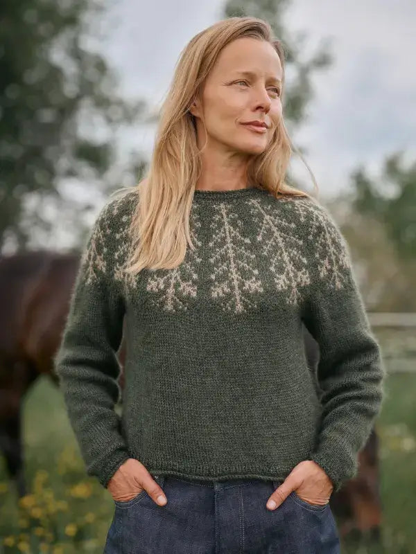 A person wears a dark green, long-sleeved Jenna Kostet - Neulottu Kalevala III, Ennakkotilaus knit sweater featuring a beige tree-pattern yoke.