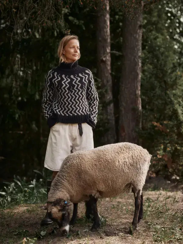 A person wearing a black and white Jenna Kostet - Neulottu Kalevala III, Ennakkotilaus sweater stands next to a sheep grazing in a forest.