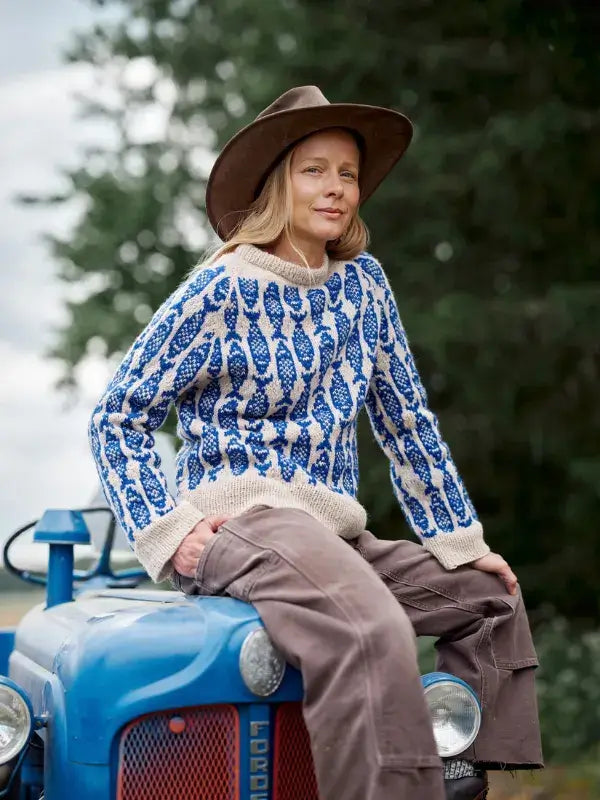 A person wearing a brown hat and a blue and beige Jenna Kostet - Neulottu Kalevala III, Ennakkotilaus knit sweater sits on the hood of a blue tractor.
