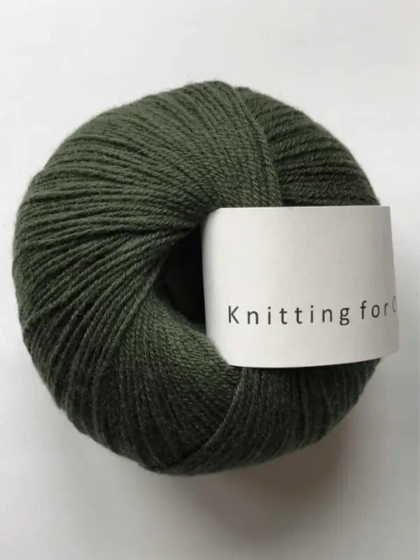 A ball of dark green Knitting for Olive Merino yarn with a white label reading "Knitting for O" rests on a plain white background. This merino wool yarn is Öko-Tex certified, ensuring extra peace of mind.