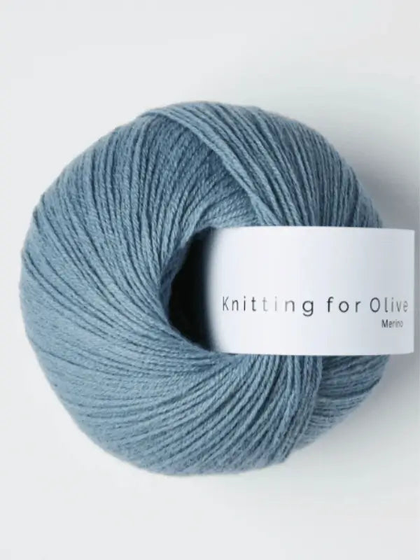 A ball of soft, blue Knitting for Olive Merino yarn with a white label rests on a plain light background. This Öko-Tex certified merino wool guarantees quality and comfort for your knitting projects.