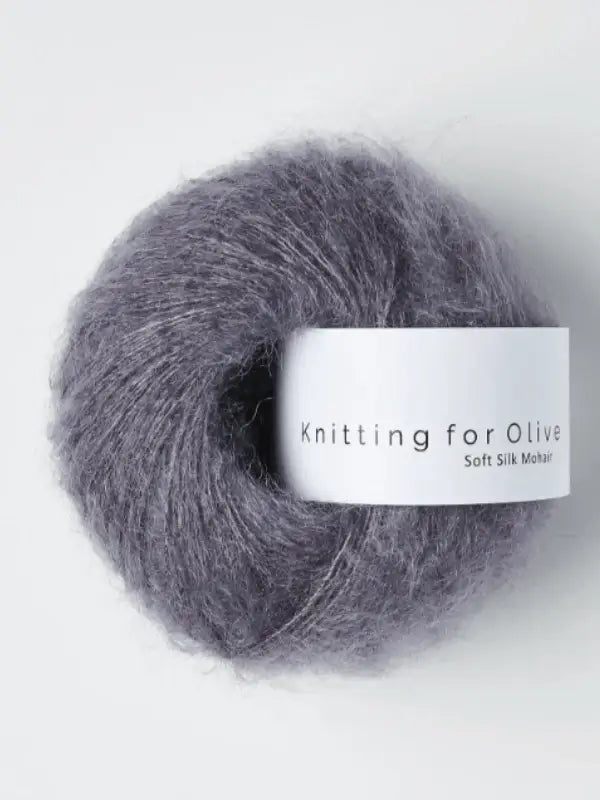 A soft, fluffy ball of purple-grey Knitting for Olive Soft Silk Mohair yarn—crafted from kid mohair and silk—features a white label displaying the brand name against a light background, exemplifying compliance with the Responsible Mohair Standard.