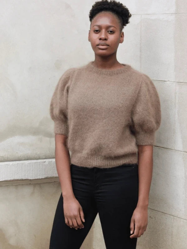 A woman stands against a light wall, wearing the brown Puff Tee neuleohje sweater with puffed sleeves and black pants. She faces the camera with a neutral expression.