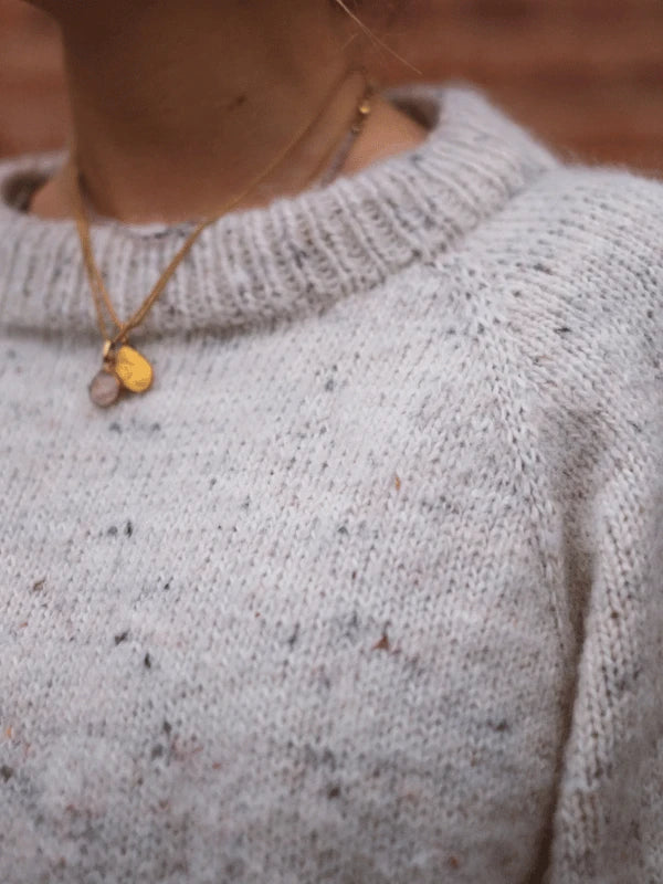 A close-up showcases a person wearing the KutovaKika Cinema Paita knit sweater in soft, light gray Sandnes Garn Sunday yarn, paired with two delicate gold necklaces featuring small round pendants. The blurred background highlights the sweater’s texture and elegant jewelry.