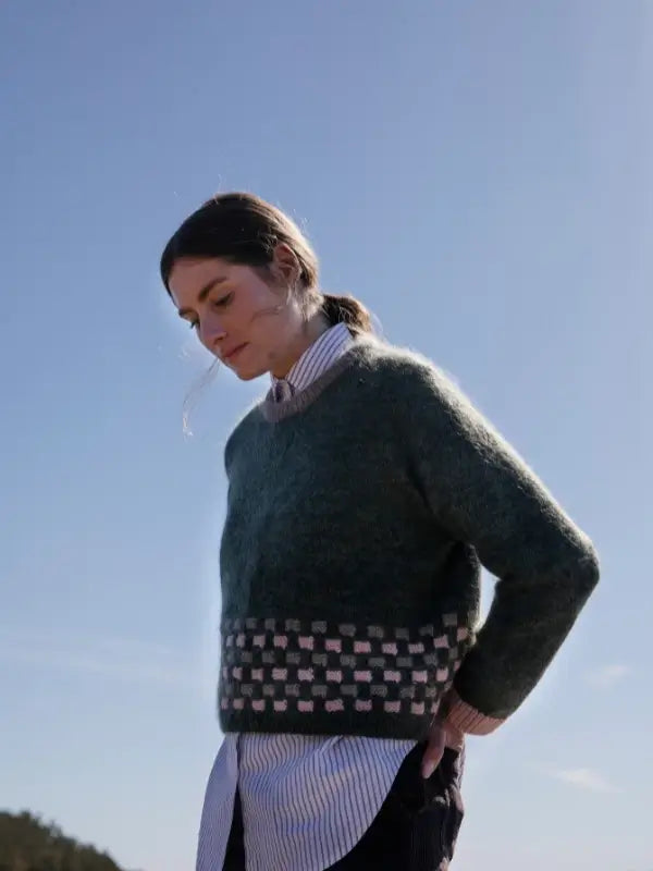 A person with long brown hair in a ponytail wears a fuzzy green checkered sweater and striped shirt outdoors under a clear blue sky, evoking the timeless northern aesthetic of Laine Juhlanumero - ENNAKKO.