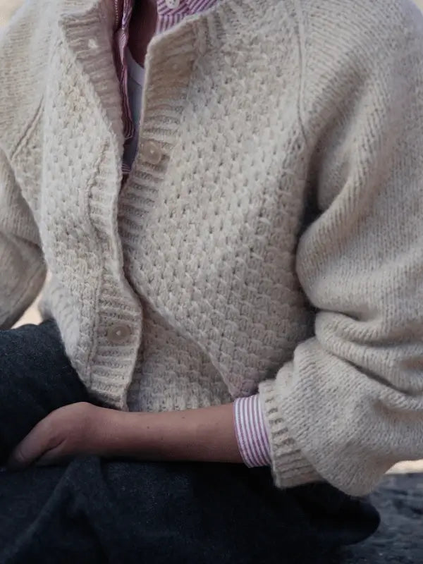 Someone wearing a textured, cream-colored Laine Juhlanumero - ENNAKKO knit cardigan inspired by pohjoismainen estetiikka over a pink and white striped shirt sits with arms crossed, shown cropped at the shoulders and waist.